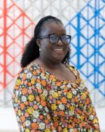 Adesola Bakare smiling, wearing glasses and a floral top, against a red, white and blue lattice backdrop.