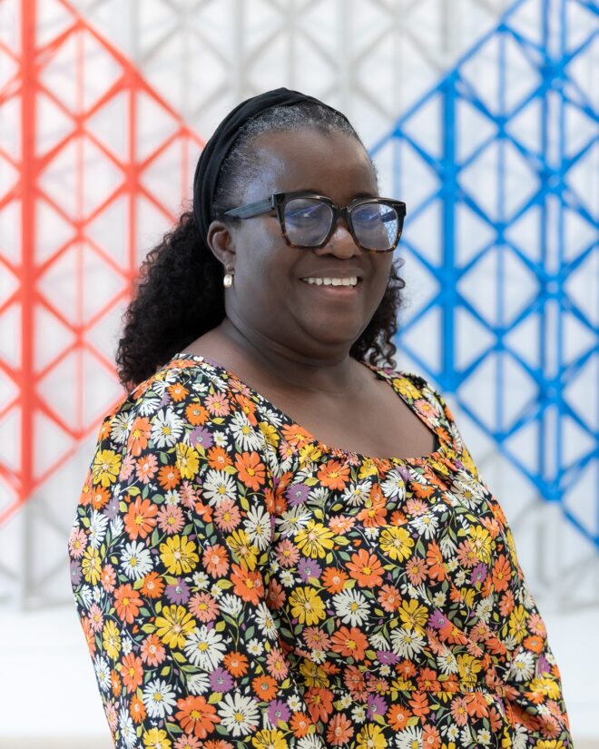 Adesola Bakare smiling, wearing glasses and a floral top, against a red, white and blue lattice backdrop.