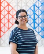 Barbie Chambers, wearing glasses and a striped navy top, standing in front of a red and blue lattice screen.