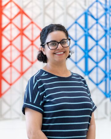 Barbie Chambers, wearing glasses and a striped navy top, standing in front of a red and blue lattice screen.