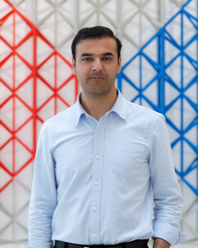 Hasan Ali Shah standing in front of a red and blue geometric lattice screen.