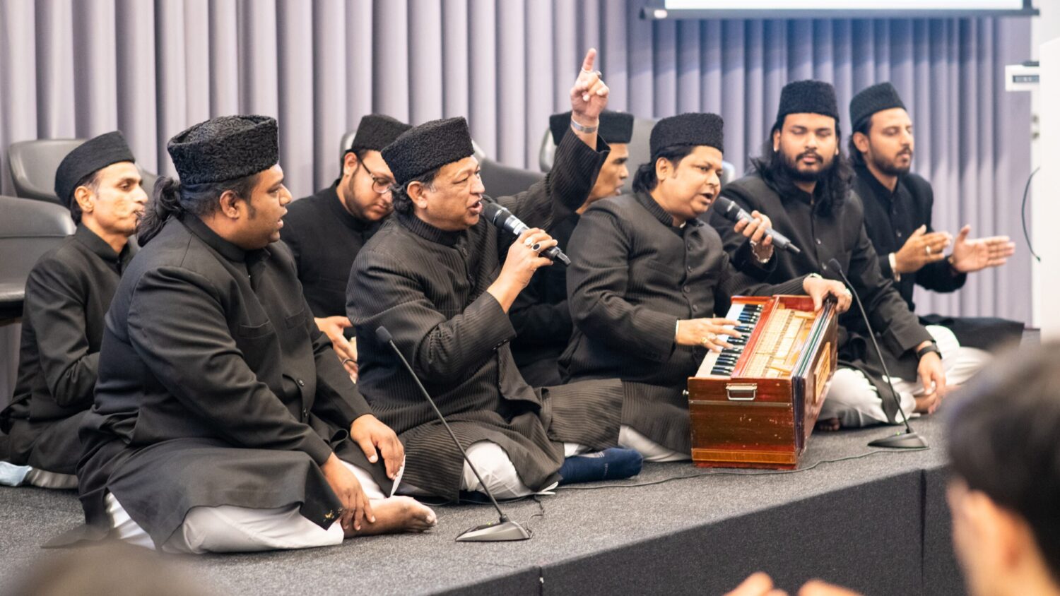 The Warsi Brothers and their qawwali group performing on stage at the Aga Khan Centre, with a harmonium in view.