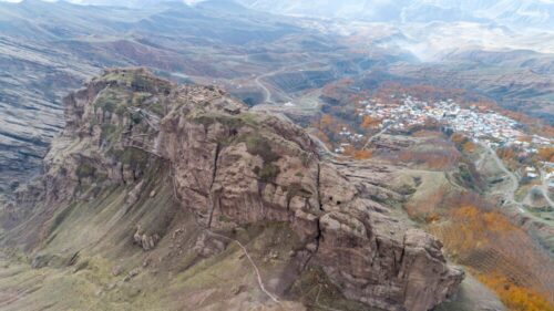 Aerial view of ruined fortress walls along a rocky mountain ridge above a valley village.