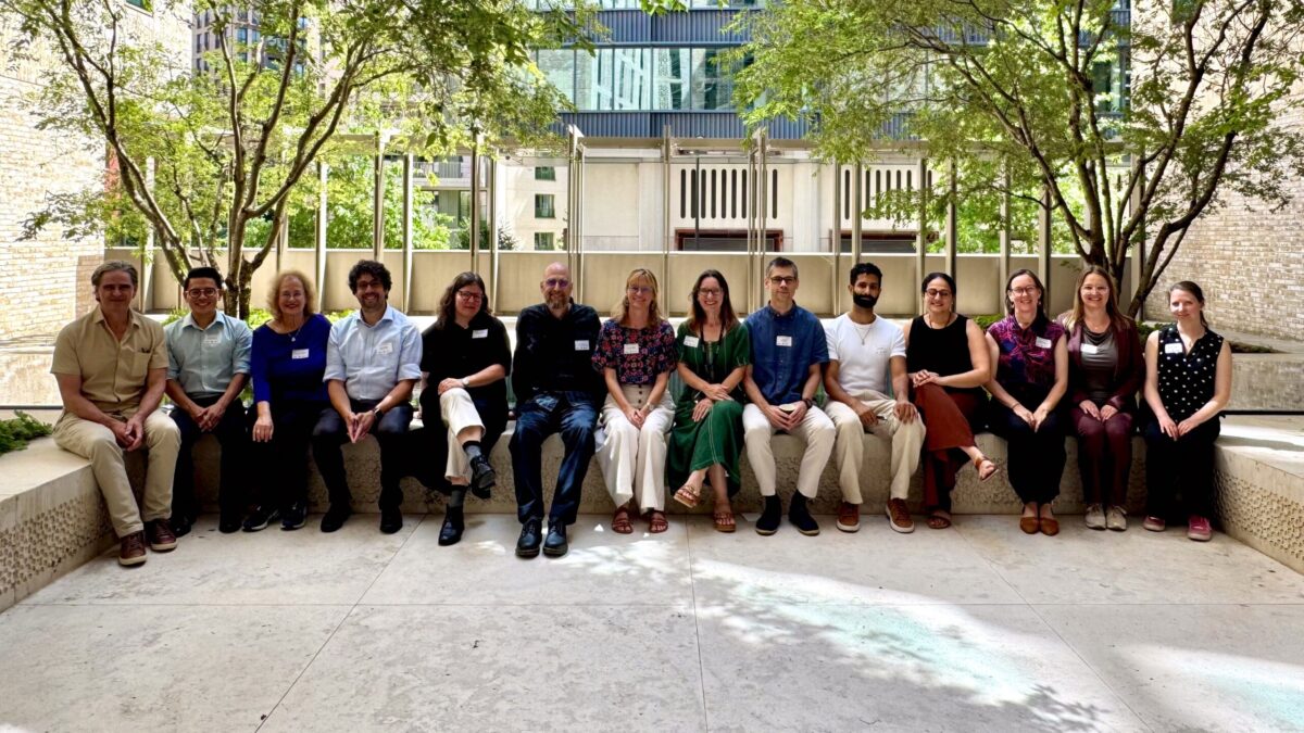 Fourteen conference participants seated together on a stone bench beneath trees in a sunlit courtyard.