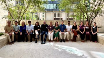Fourteen conference participants seated together on a stone bench beneath trees in a sunlit courtyard.