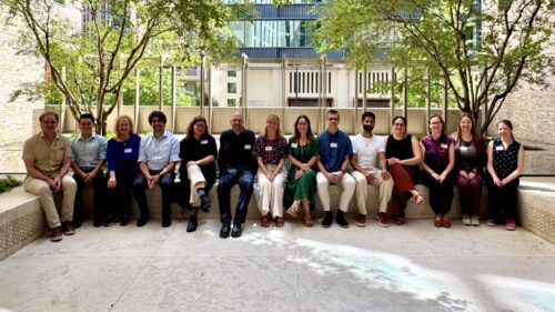 Fourteen conference participants seated together on a stone bench beneath trees in a sunlit courtyard.