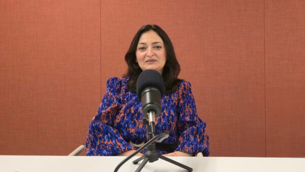 Qudsia Shah seated at a table speaking into a studio microphone during a podcast recording.