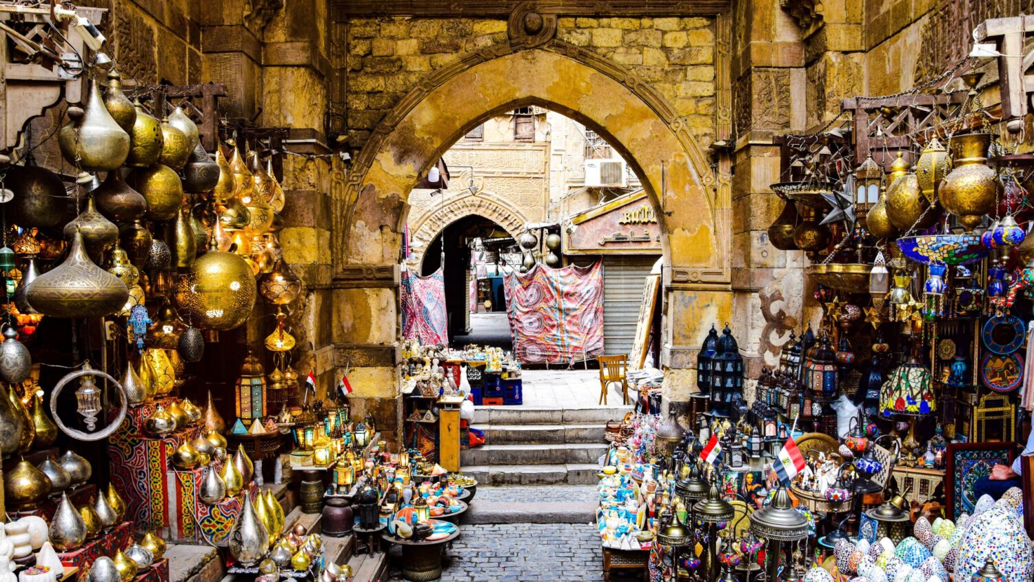 Brass and glass lanterns hanging around a stone archway leading into a covered market lane in Khan al-Khalili, Cairo.