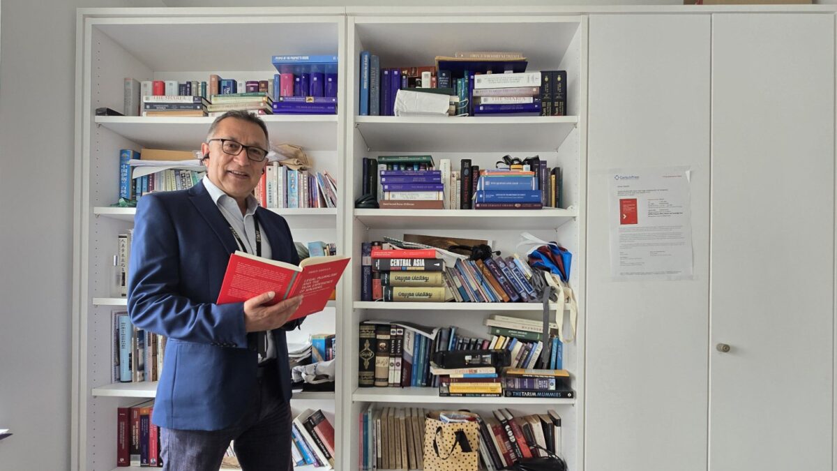 Dr Amier Saidula holding his new book on legal pluralism among the Tajiks of Xinjiang, in front of academic bookshelves.