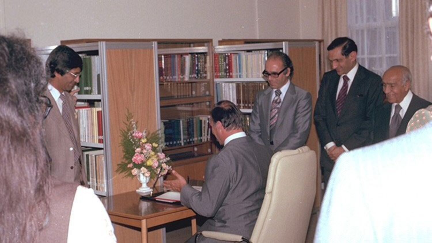 Archival photograph in memorium of a seated man signing a book at a table, watched by Jalal Badakhchani in a library.