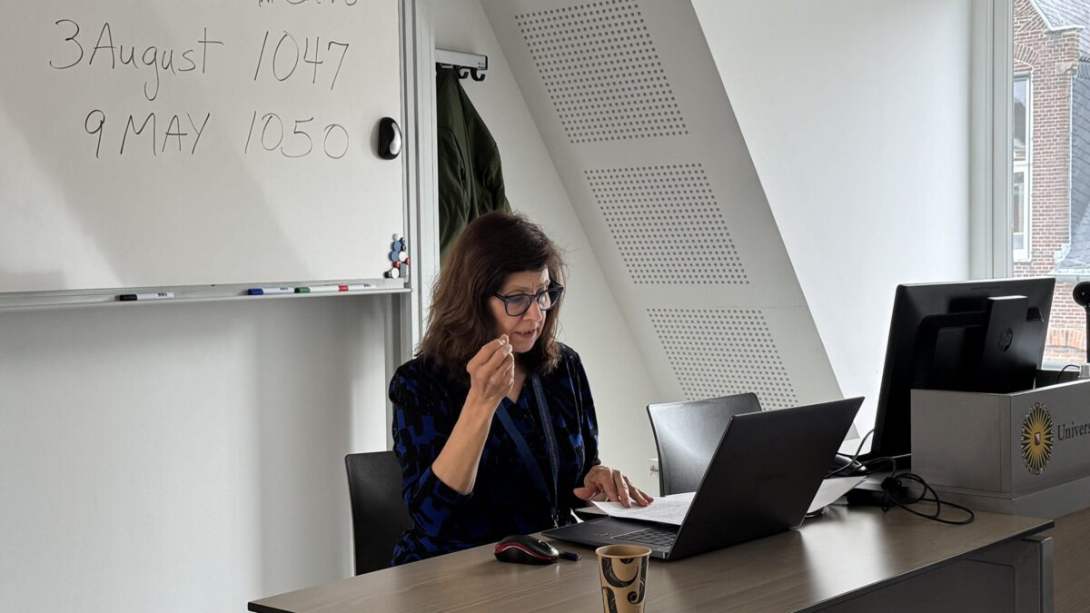 Dr Alice Hunsberger speaking at a desk, notes and laptop in front of her, whiteboard behind