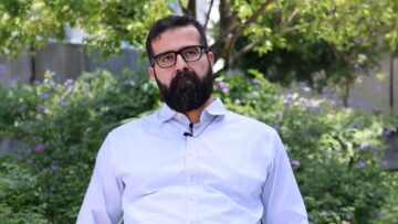 Dr Hasan Al-Khoee speaking to camera with a lapel microphone, seated outdoors in front of a flowering garden.