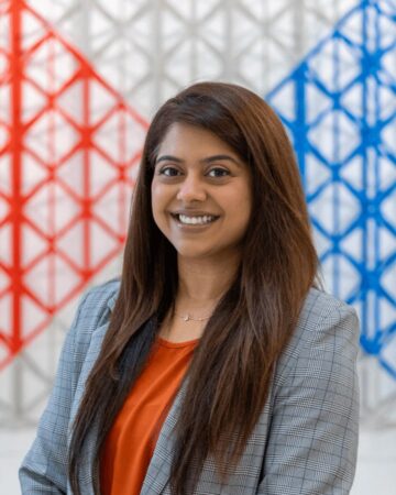 Heena Jiwani smiling, wearing a grey checked blazer, in front of a red and blue geometric lattice installation.