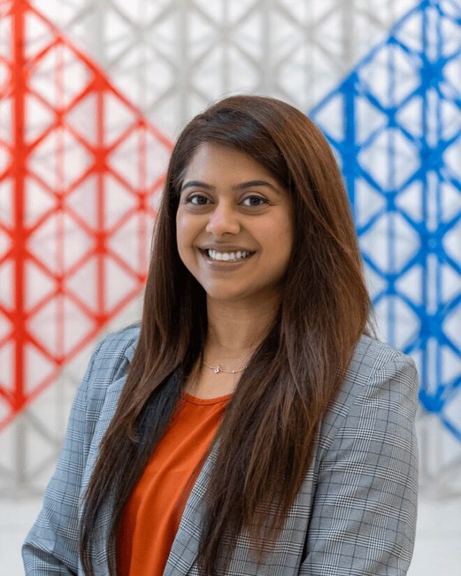 Heena Jiwani smiling, wearing a grey checked blazer, in front of a red and blue geometric lattice installation.