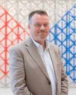 Oliver Scharbrodt, wearing a beige blazer and pale blue shirt, in front of a red, white and blue geometric lattice.
