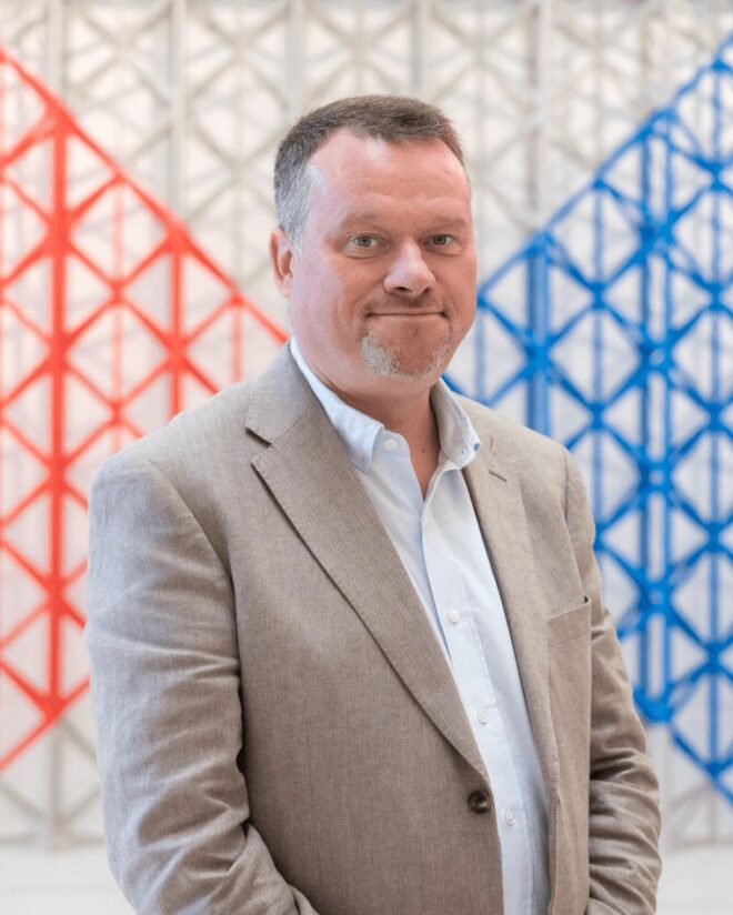 Oliver Scharbrodt, wearing a beige blazer and pale blue shirt, in front of a red, white and blue geometric lattice.