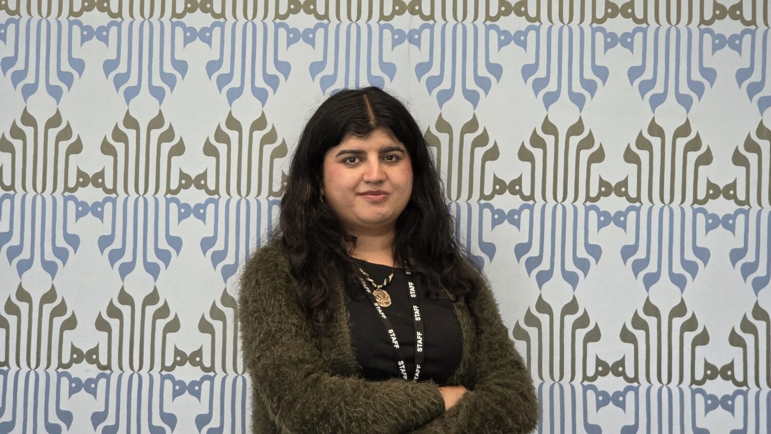 Dr Zehra Kazmi, arms folded, standing in front of a wall decorated with a blue and olive calligraphic pattern.
