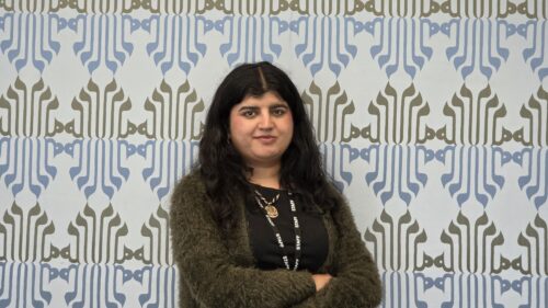 Dr Zehra Kazmi, arms folded, standing in front of a wall decorated with a blue and olive calligraphic pattern.