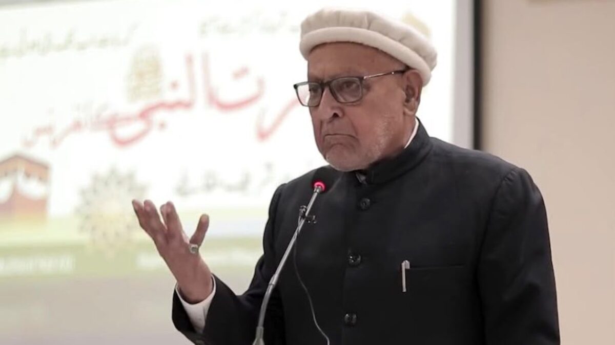 Alwaez Fida Ali Aisar in a white wool cap and black coat, speaking at a microphone with one hand raised.