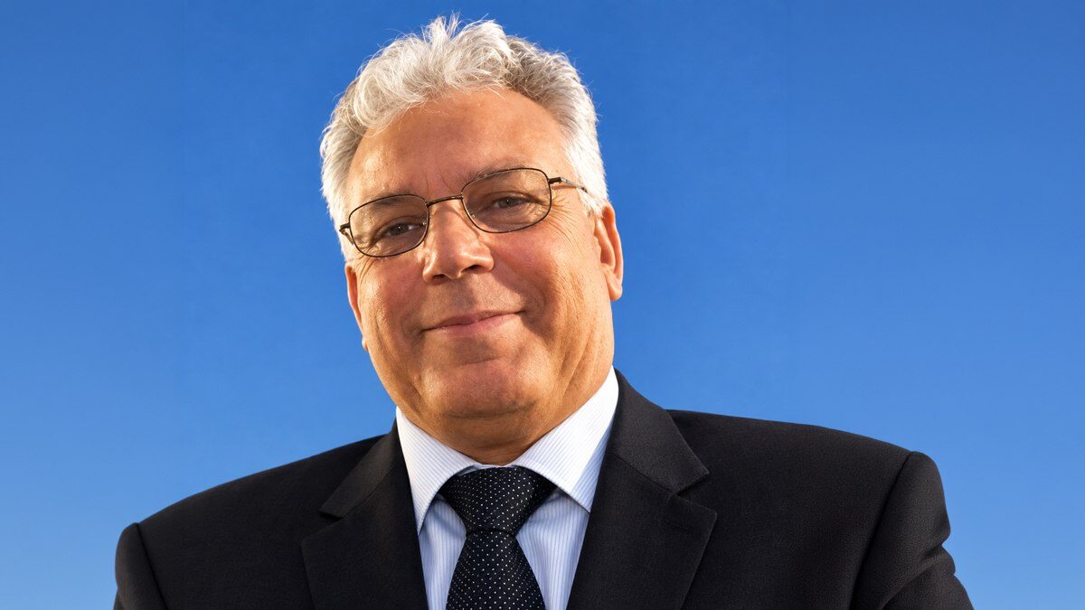 Ali Suhail Kabalan in a dark suit and glasses, smiling, photographed against a clear blue sky.