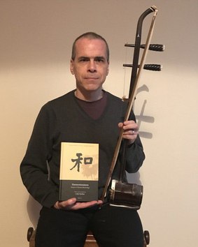 Colin Huehns holding a two-string bowed fiddle and a book with a Chinese character on its cover.
