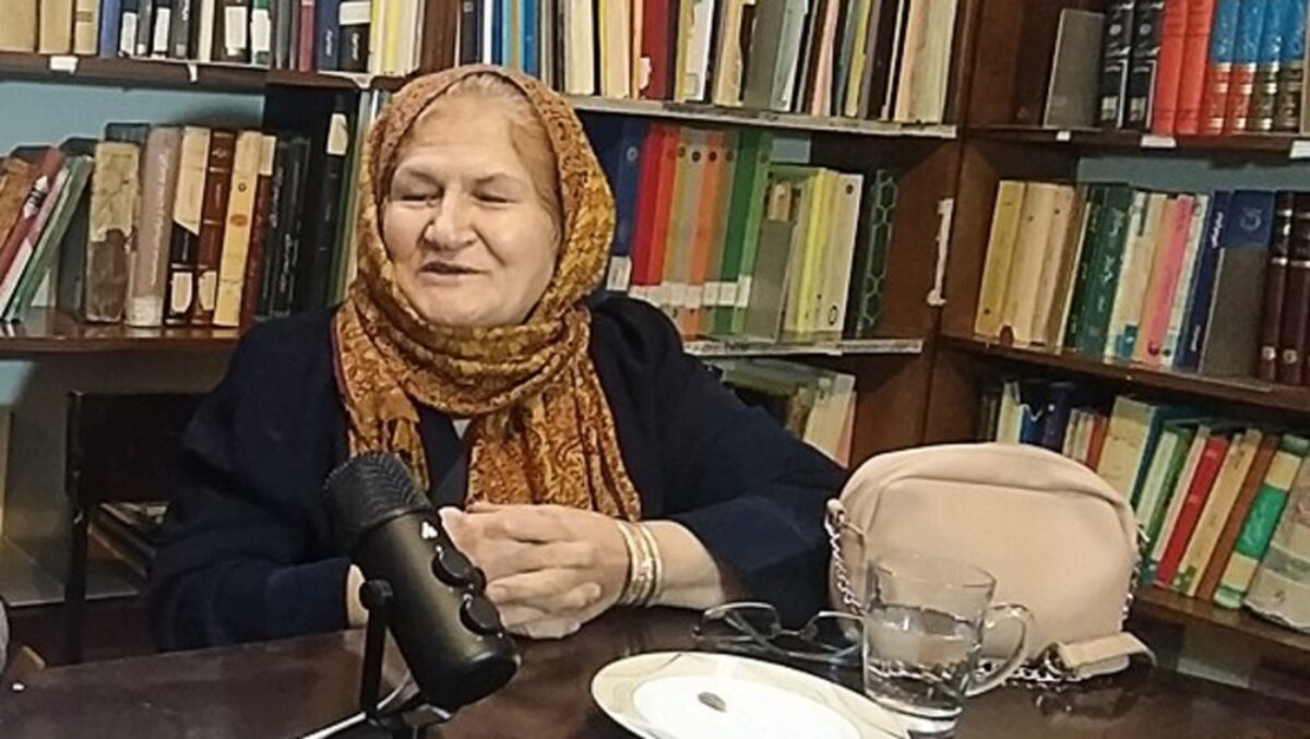 Fatemeh Nisa Mirshahi seated at a table with a microphone in front of her and bookshelves behind.