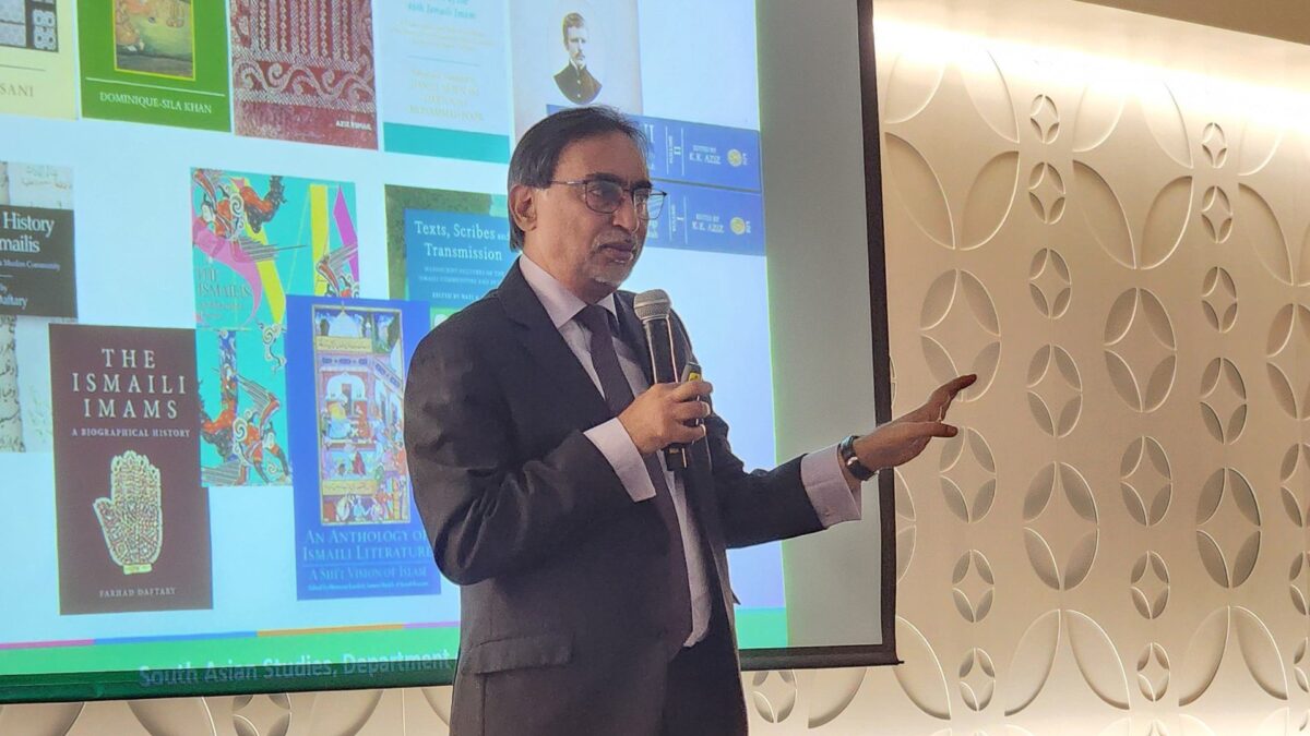 Hussain Jasani speaking into a microphone and gesturing beside a screen displaying IIS book covers.
