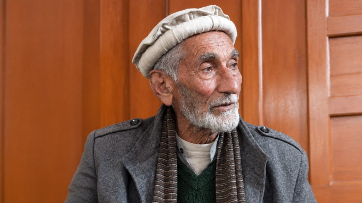 Muhammad Munir Khan in a cream wool cap, grey coat and striped scarf, looking to one side.