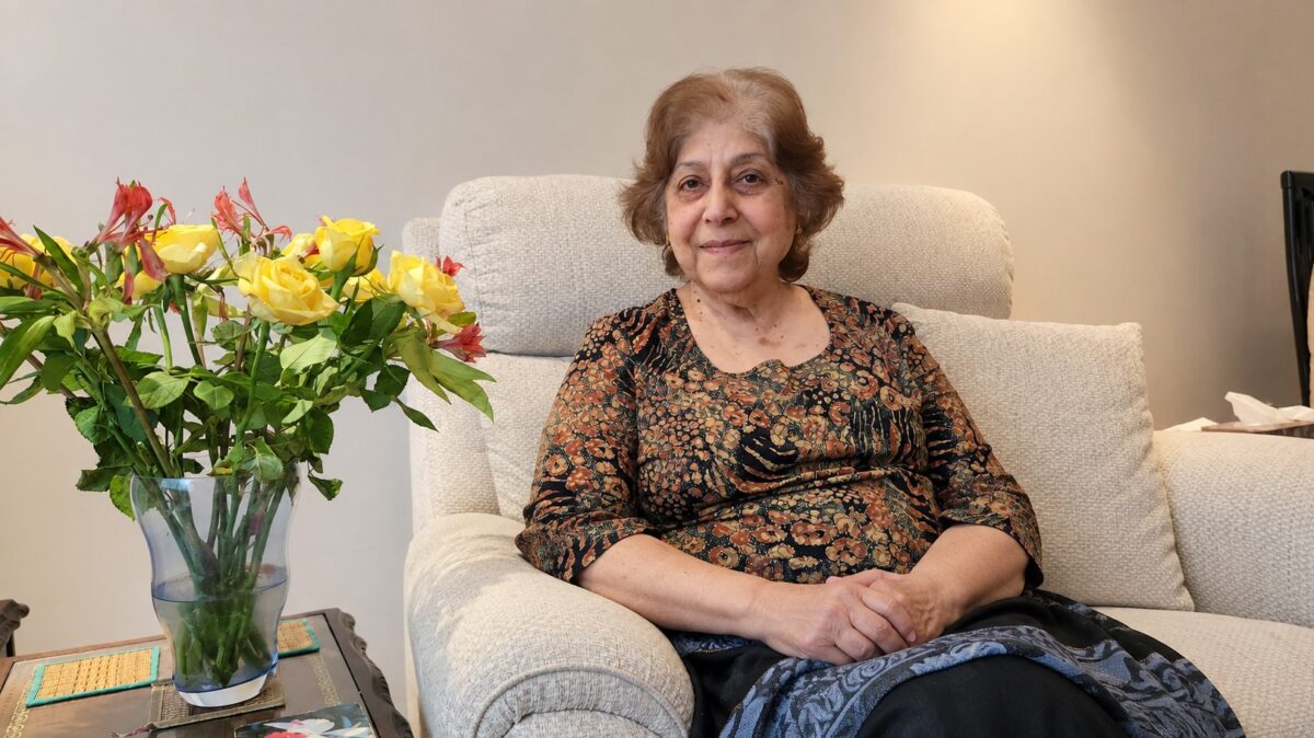 Zubeda Firoz Bhagat seated in a cream armchair beside a vase of yellow roses, smiling at the camera.