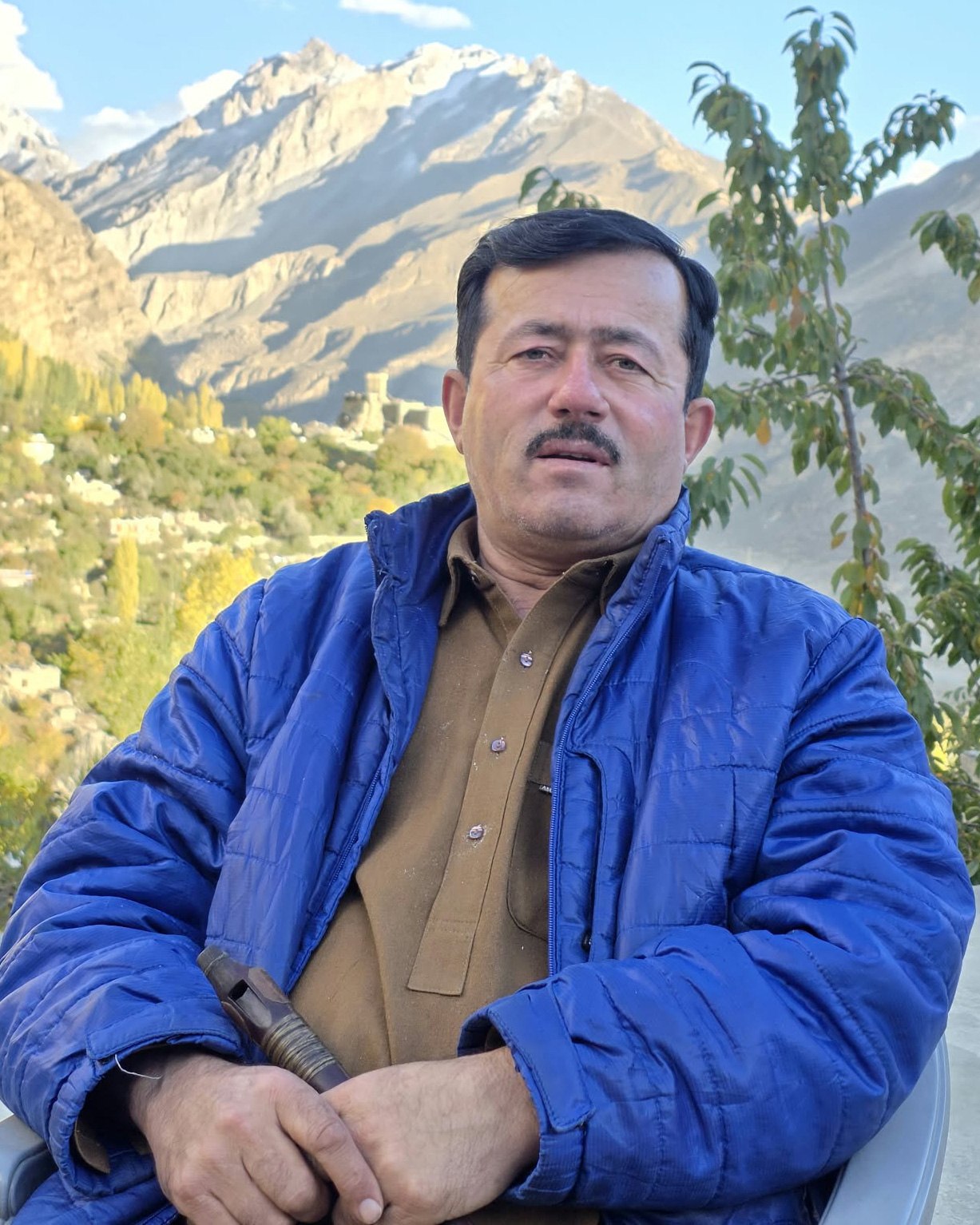 Ustad Abdul Karim seated outdoors holding a small wooden wind instrument, with a mountain valley behind.