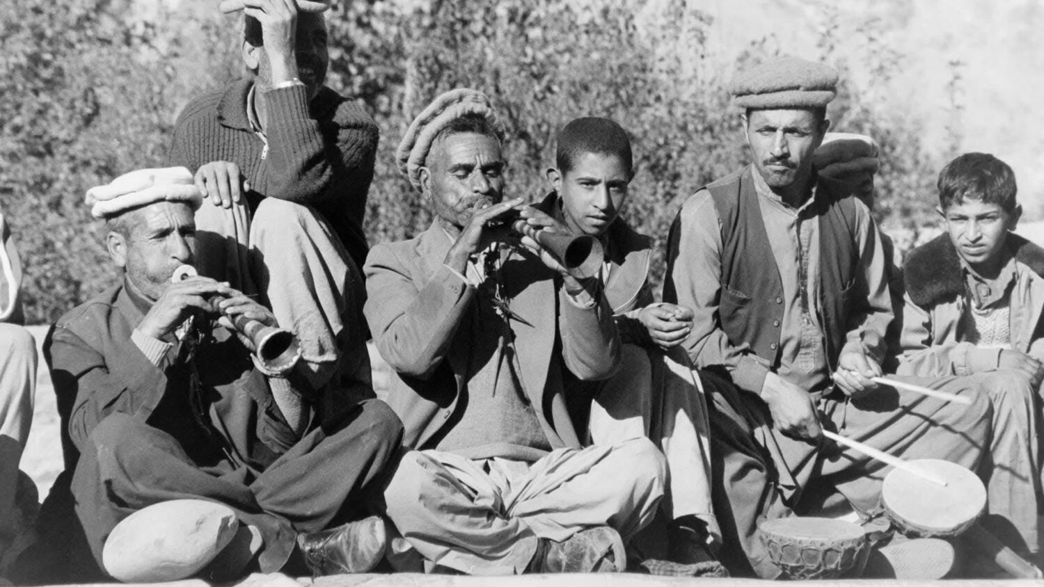 Black-and-white photograph of seated musicians playing wind instruments and drums, with onlookers.
