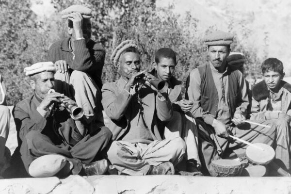 Black-and-white photograph of seated musicians playing wind instruments and drums, with onlookers.