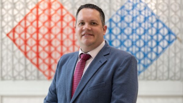 Dr Stephen Burge, smiling, in a blue blazer and red tie, standing before a red and blue geometric lattice artwork.