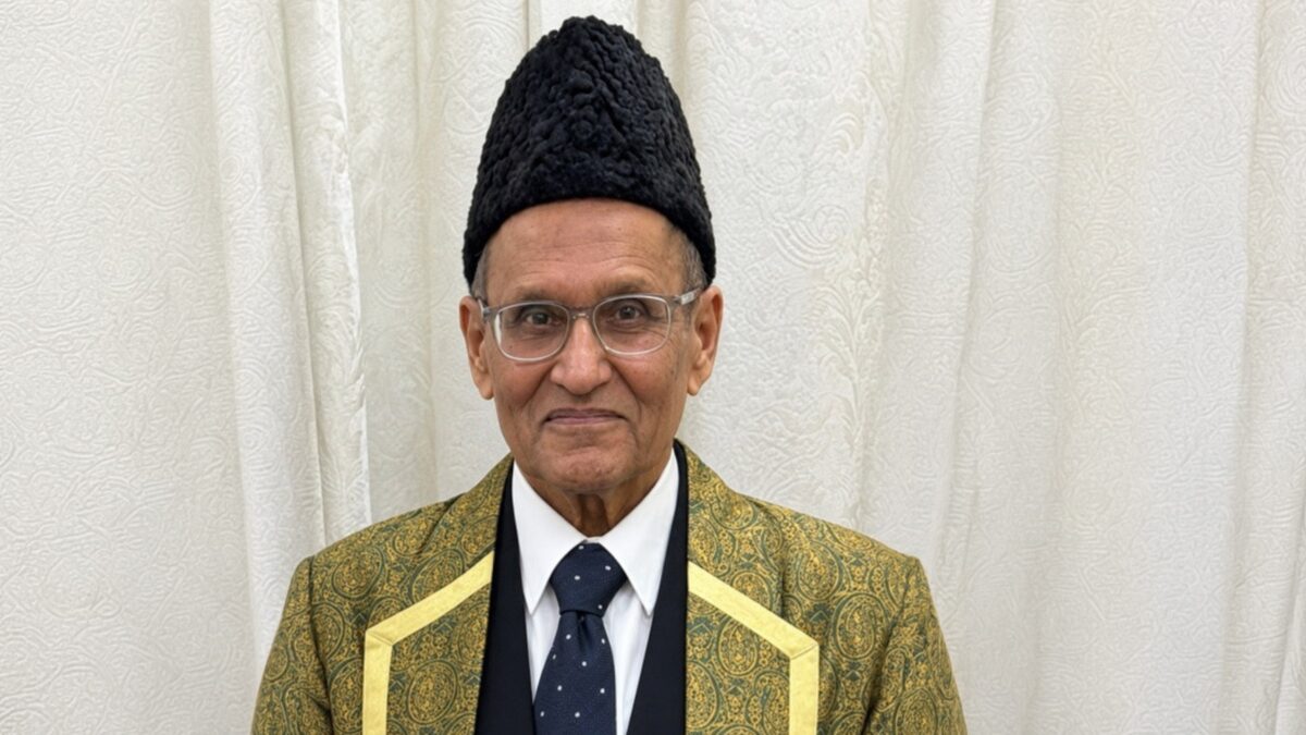 Karim Ratansi in a black cap and a green and gold patterned robe over a dark suit, smiling at the camera.