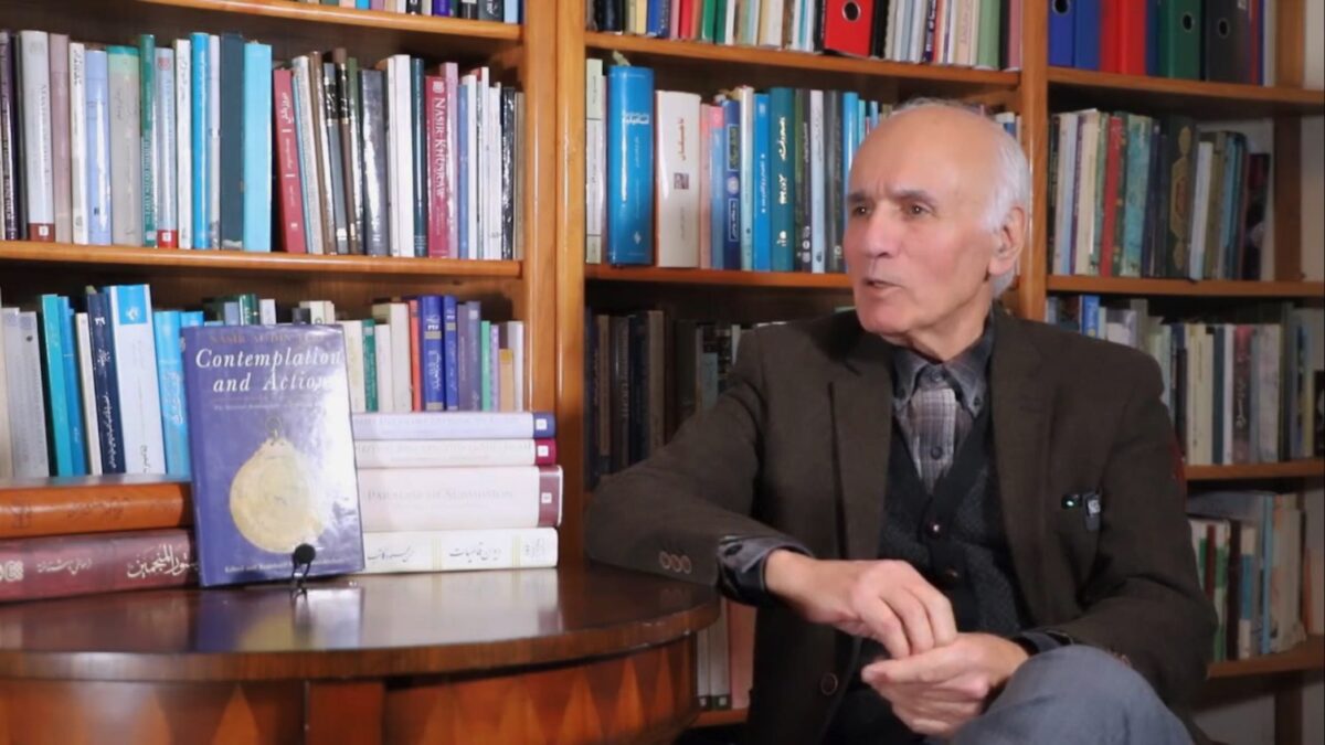 Dr Jalal Badakhchani seated beside a table of books in a book-lined room, speaking and gesturing.